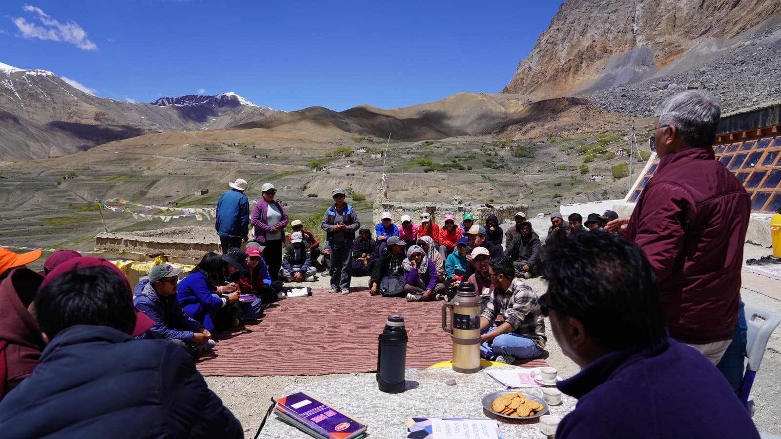 The monitoring team and community members seated together for a consultation on a terrace, mountains behind