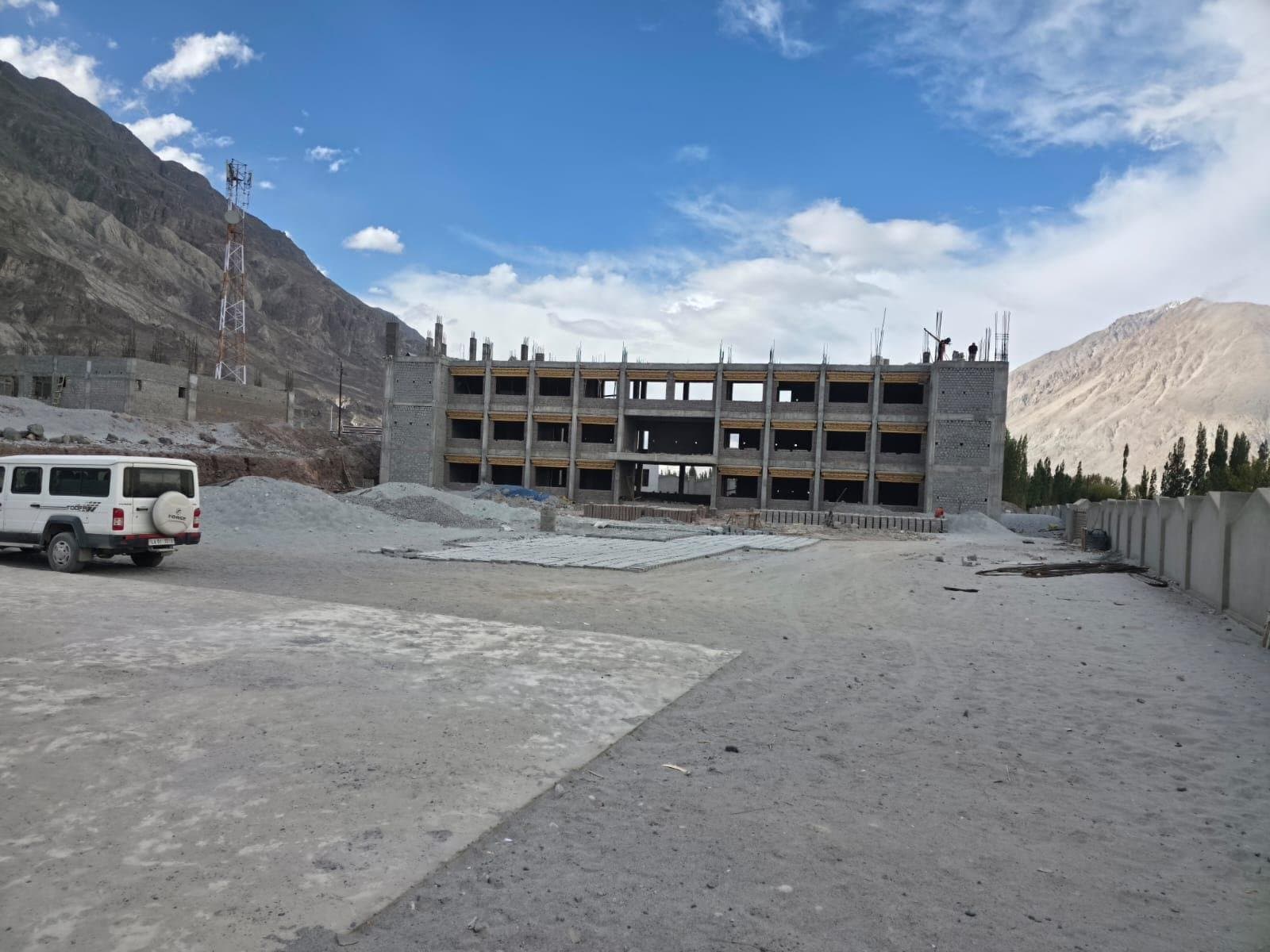 A new multi-storey school building under construction beside a cleared ground, mountains behind