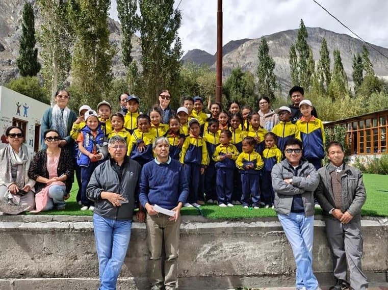 The DIET Leh monitoring team with students in yellow and blue uniforms and their teachers on a school lawn, poplar trees and mountains behind