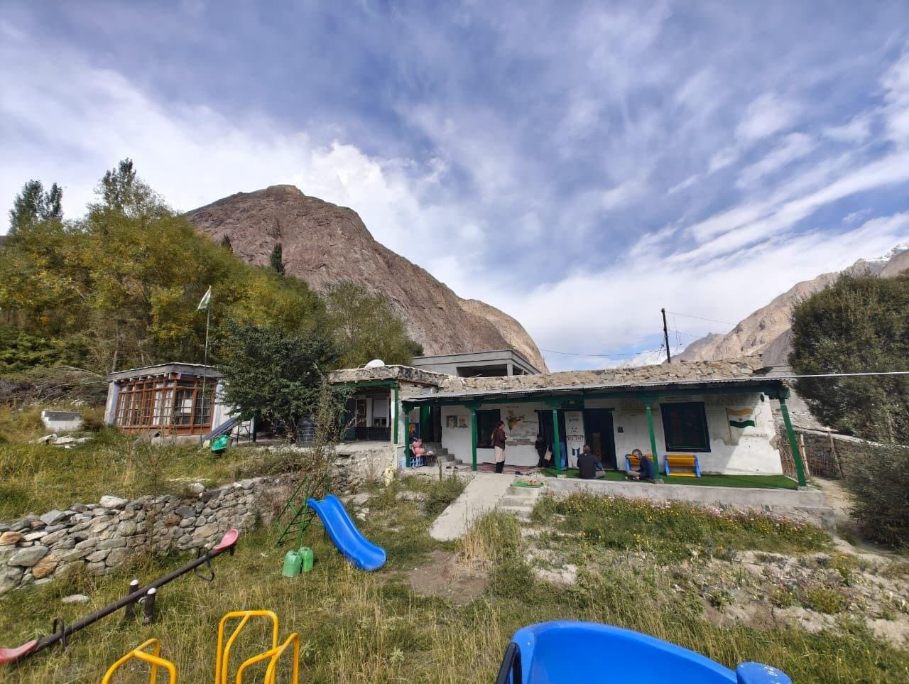 A small single-storey village school with a slide and play equipment on the grass in front, under a wide sky