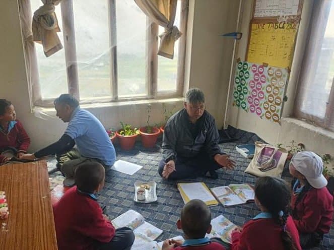 Members of the monitoring team seated on the floor with a group of students reading from their books