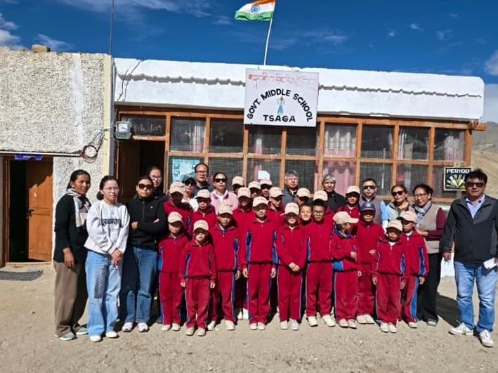 The monitoring team with students in red uniforms and staff outside Govt. Middle School, Tsaga, beneath the national flag