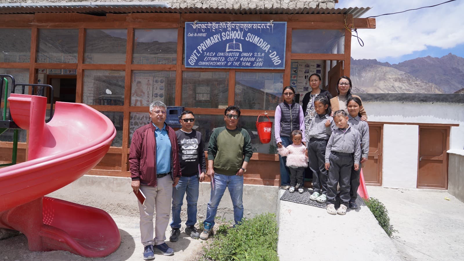The monitoring team with students and staff outside Govt. Primary School, Sumdha-Dho, beside a playground slide