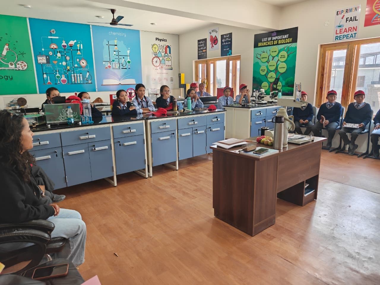 School students in uniform seated at the lab benches and along the wall during a session, with a teacher seated in the foreground