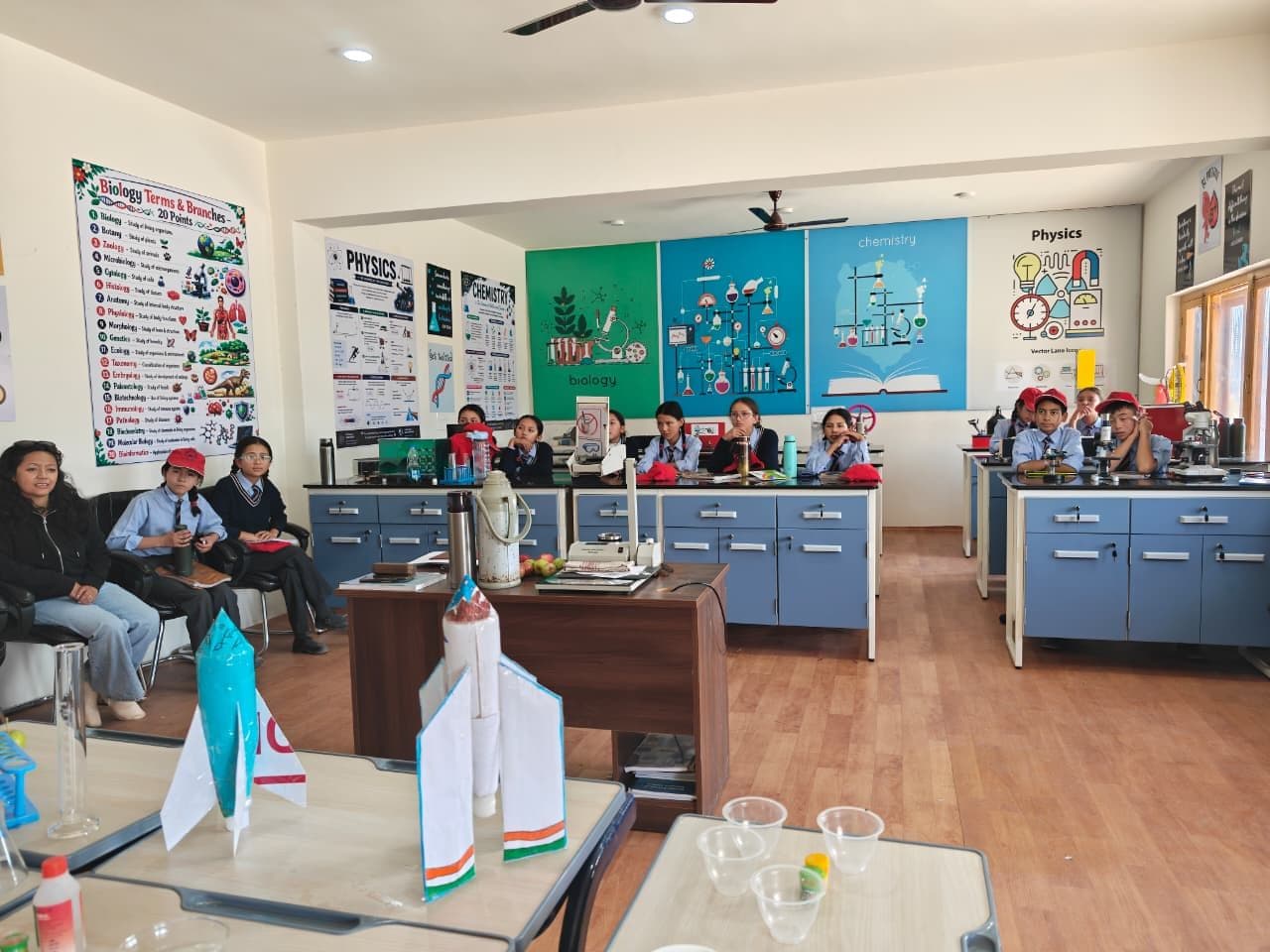 School students in uniform seated at lab benches during a visit to the science laboratory, with model rockets in the foreground