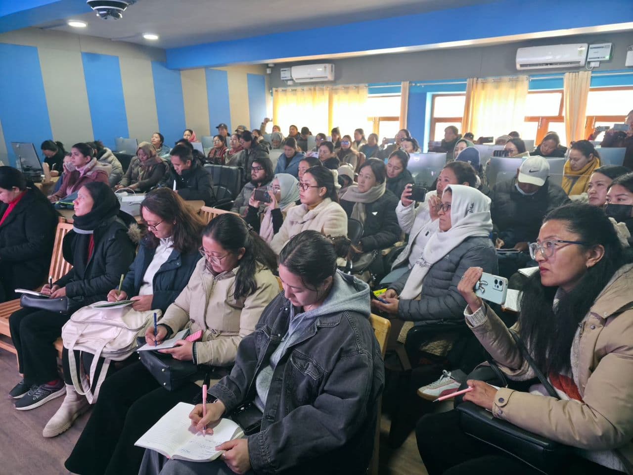 Teachers seated in the Vidya Samiksha Kendra hall, several taking notes, during a session